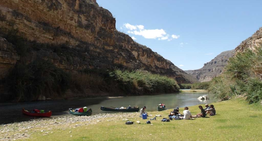 A group of people rest on the green shore of a river framed by tall canyon walls. Canoes rest on the beach nearby. 